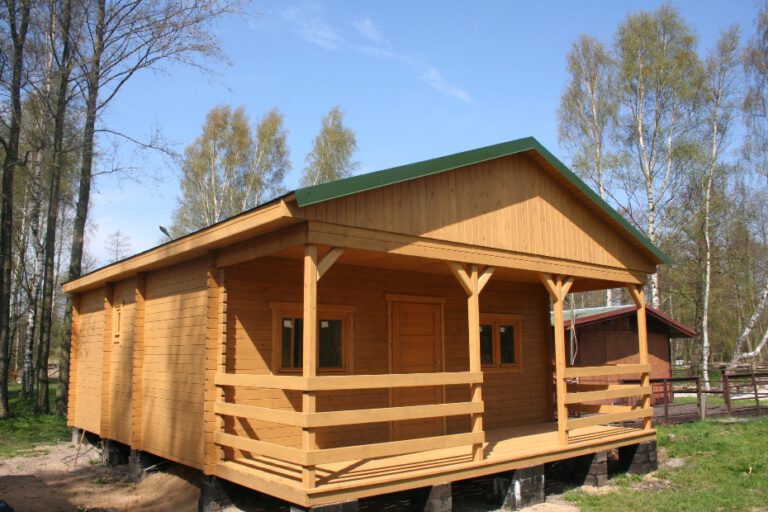 Drewniany domek letniskowy z zadaszonym tarasem, otoczony zielenią.